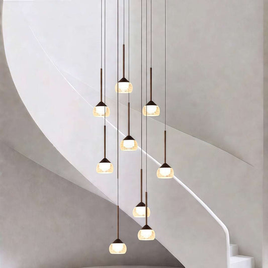 A modern multi-light pendant chandelier with nine glowing glass shades and dark metal accents, suspended against a minimalist curved gray wall and staircase.